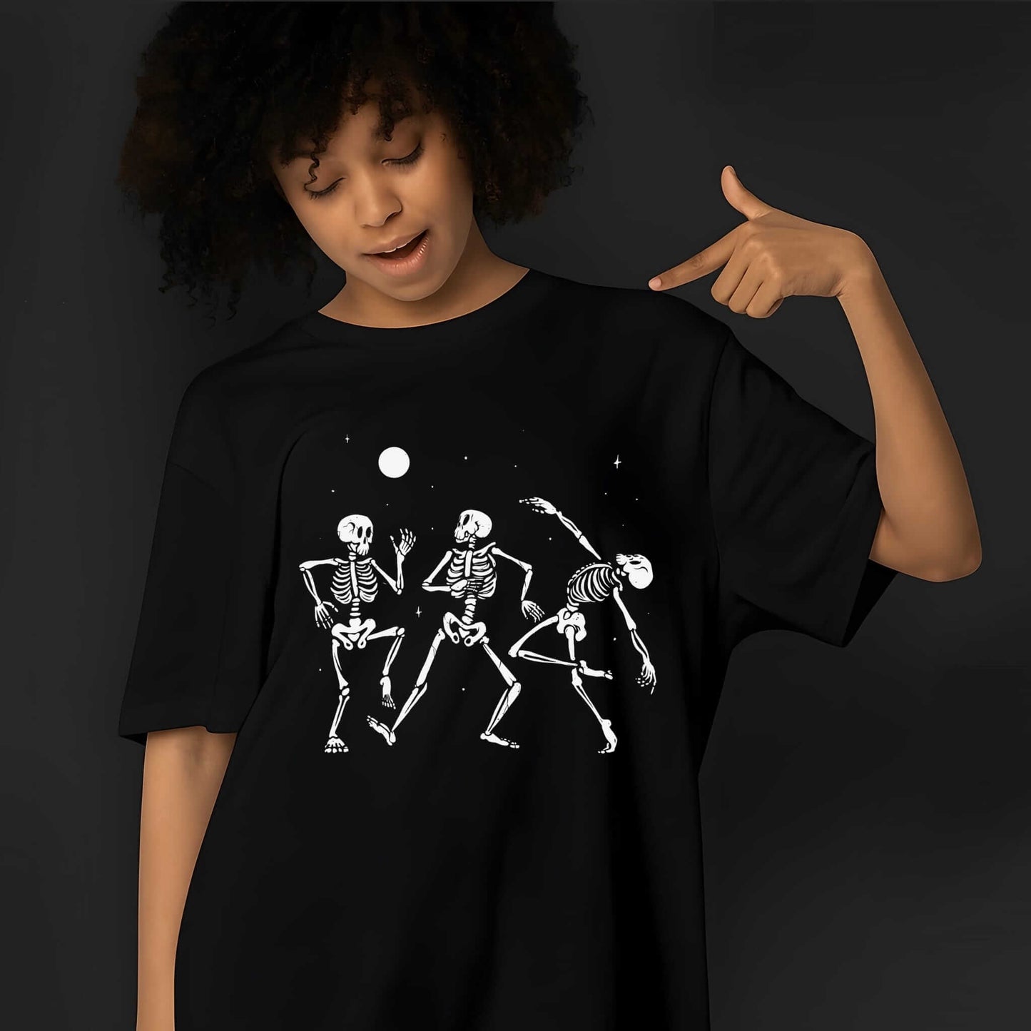 Three white skeleton figures dancing together under a full moon and stars in playful, animated poses on a black t-shirt.