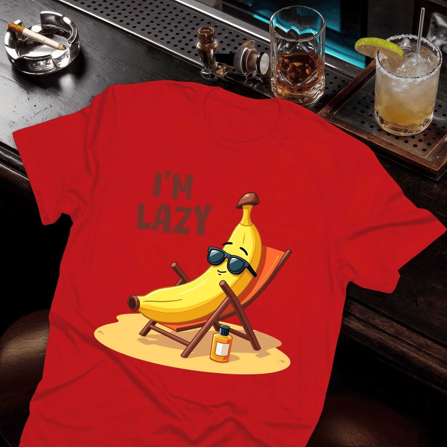 Red t-shirt "I'M LAZY" text with relaxed banana in sunglasses lounging on beach chair and sunscreen on sandy patch