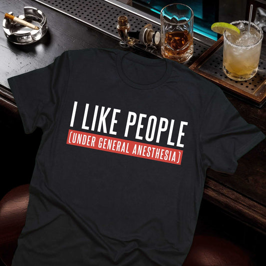 Black tee with witty medical humor text "I LIKE PEOPLE (UNDER GENERAL ANESTHESIA)" in contrasting light typography