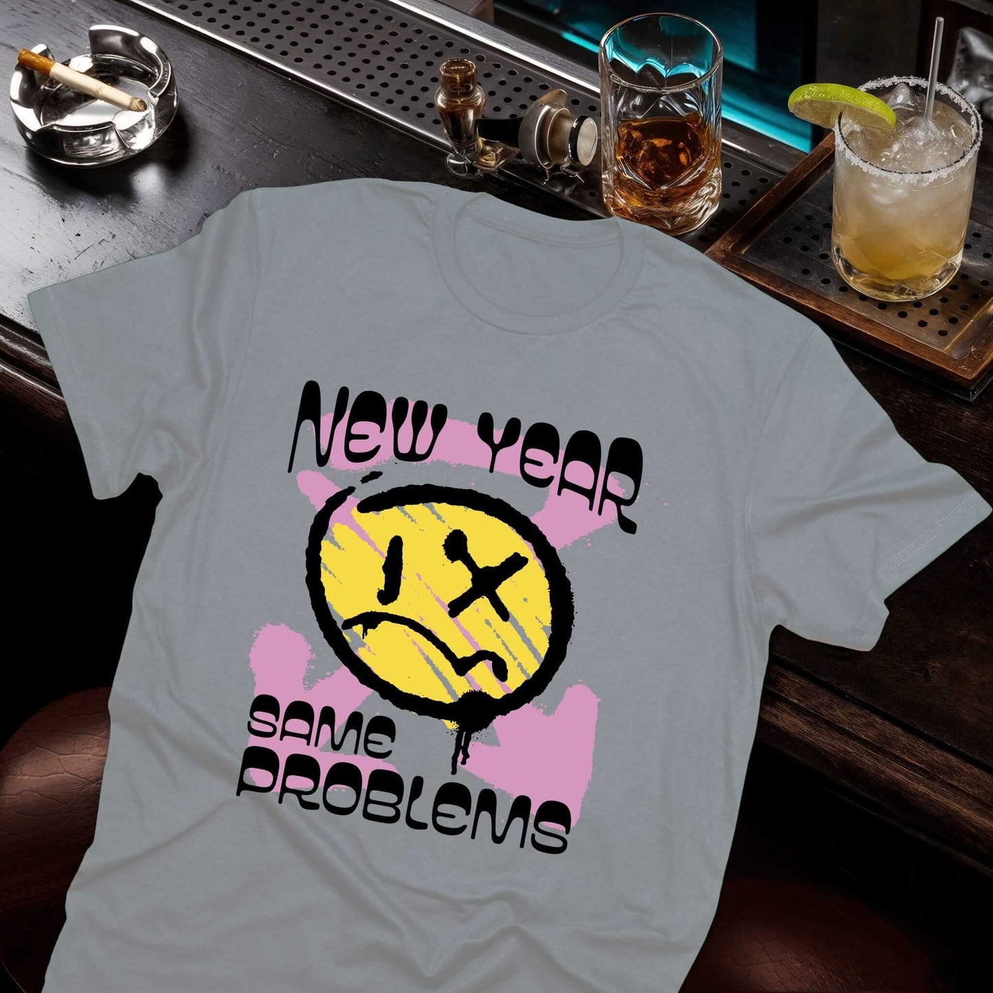 Funny sarcastic face with X eye, colorful paint splashes, and humorous "New Year Same Problems" text design on gray t-shirt.