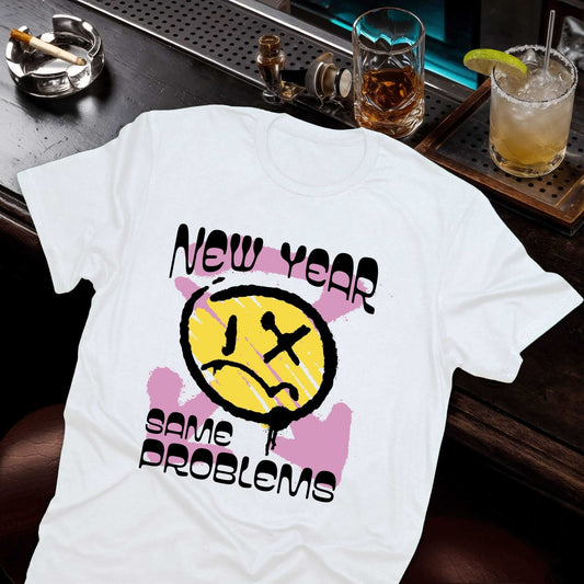 New Year Same Problems design with yellow smiley face, X-ed out eye, pink splatter effects, and bold black text on white tee.